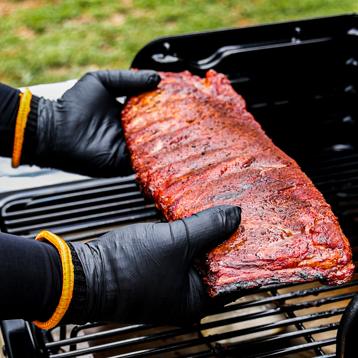Black-gloved hands lifting a rack of barbecued ribs from the grill