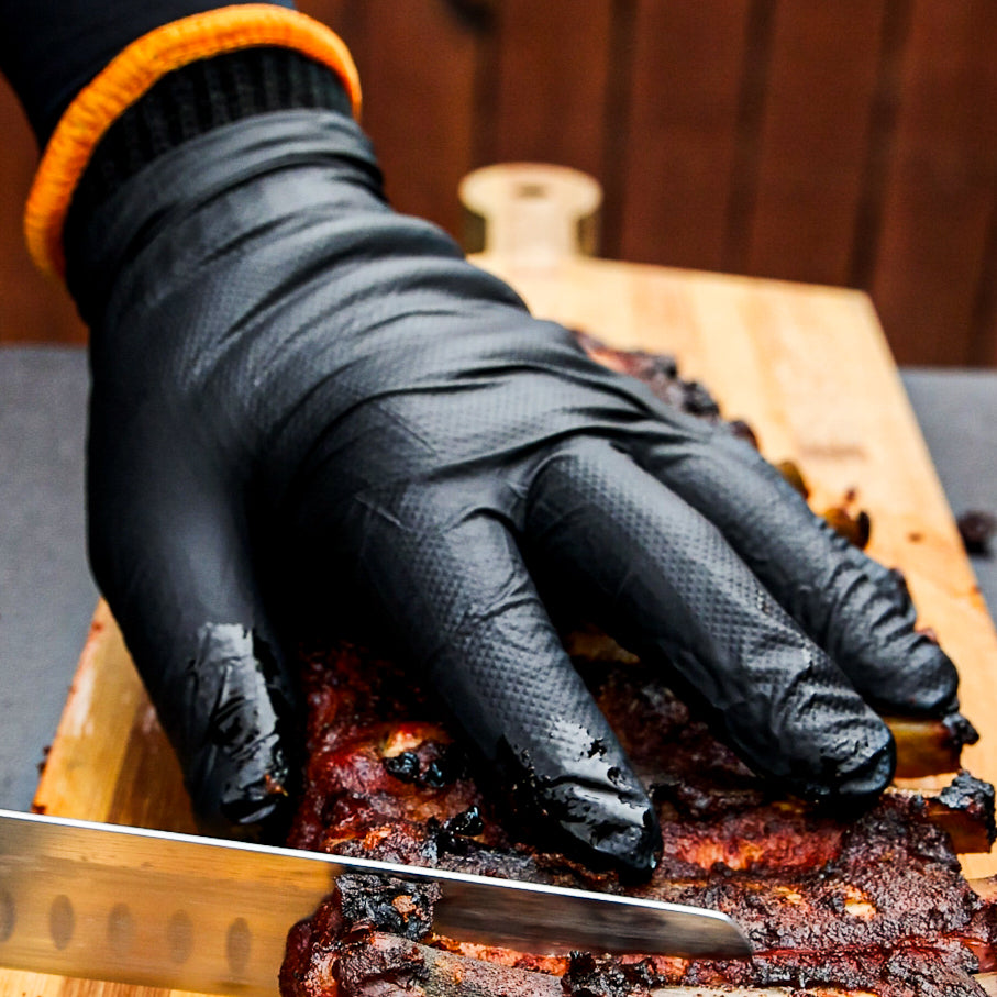 A gloved hand holding barbecued ribs steady while slicing on a cutting board